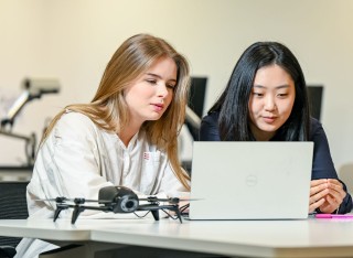 Two young women work together on a computer with a drone alongside