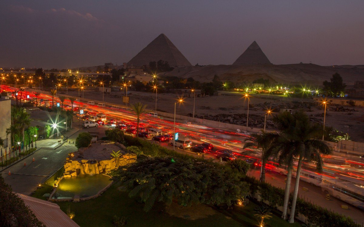 Time lapse photo showing the lights from moving traffic along a busy Cairo street with pyramids silhouetted against the skyline in the distance