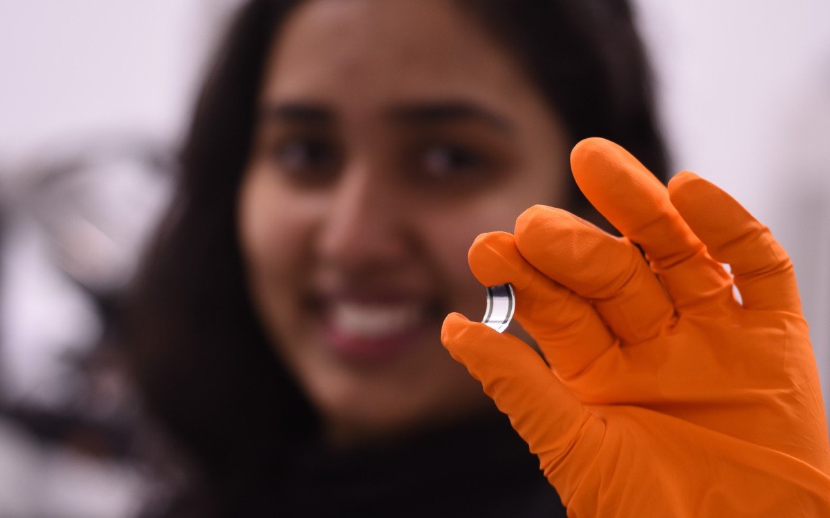 A blurred face smiles behind a hand, clad in an orange lab glove, holds a tiny rectangle of the new x-ray