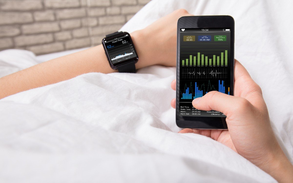 Two hands of a person lying in a bed with crisp white sheets. The left wrist wears a smart watch showing health data. The right hand holds a mobile phone