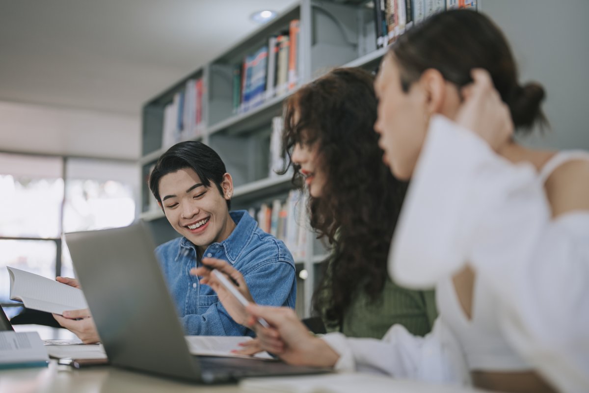 3 Asian LGBTQ+ adult students studying in college library writing notes and using laptop discussion on homework school project