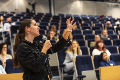 A lecturer talks with a microphone to a room of students. 