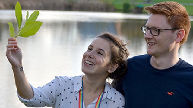 Two students are standing in front of the campus lake, one student is holding up a leaf which they are both looking at