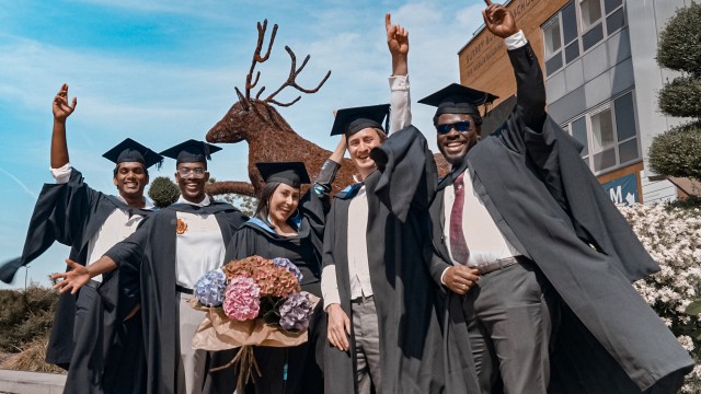 A group of postgraduate students at graduation cheering in front of a blue sky.