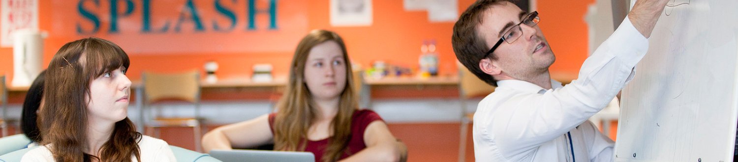 A teacher is writing on a white board while two students look on.