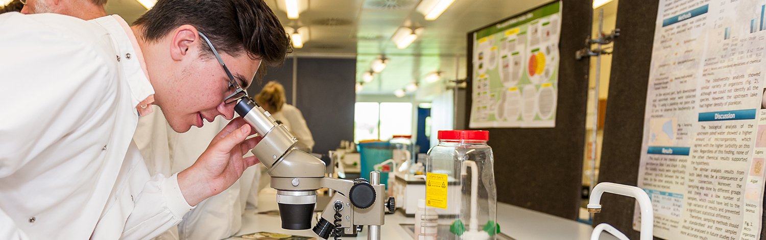 A student is wearing a white lab coat and looking into a microscope