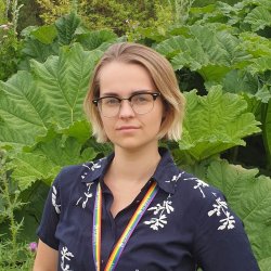 An adult with short blond hair, glasses and a rainbow lanyard, standing in front of large green foiliage