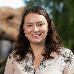 Michelle, a white woman with brown hair, stands in front of a rollercoaster she designed, smiling at the camera.