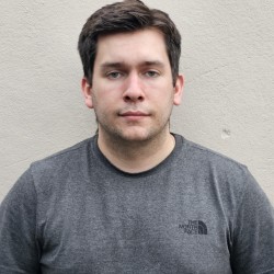 Ryan, a white man with brown hair wearing a grey t-shirt, looks at the camera in front of a white wall.