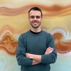 Charlie is a white man with short dark hair, he is wearing a green jumper and smiles at the camera with his arms crossed.