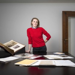A picture of Katherine with some archive materials. She is standing wearing a red jumper. 
