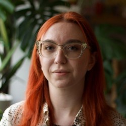 The picture portrays a young white woman with orange hair. The picture is a close-up from the neck up. Background includes some blurred plants.