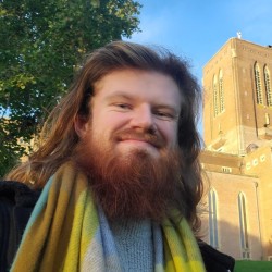 Photo of Adam Gorry smiling at the camera, with a sunset-lit Guildford Cathedal grounds behind him.