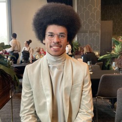 The image shows an afro carribean and white male in a velvet jacket along with a velvet neck tight shirt. He smiles at the camera and has a large afro and wears glasses. He is sitting at a table and there is an aura of happiness about him. His name is Lloyd La Ronde.