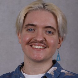 An image of a person smiling at the camera. He is white, with bleached blonde hair and a moustache. The image shows his shoulders and head. He has a blue shirt with a white bird pattern and a matching blue earring. 