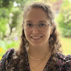 White female with brown curly hair and round glasses. Facing forwards smiling at the camera. Green blurred garden background.