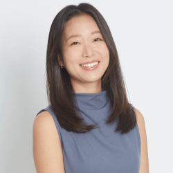 Melanie Hyo-In Han is smiling at the camera against a light grey background. She has shoulder-length dark brown hair and is wearing a slate-blue top. 