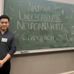 Michael stands in front of a blackboard that says 'National Undergraduate Neuroanatomy Competition'.