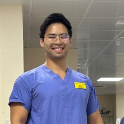Wei Chin smiles at the camera wearing blue scrubs in his part-time healthcare role.