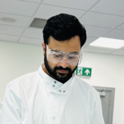 Aagaz wears PPE in the lab and is looking down at an experiment.