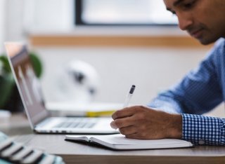 A young man writing on a piece of paper next to an open laptop.