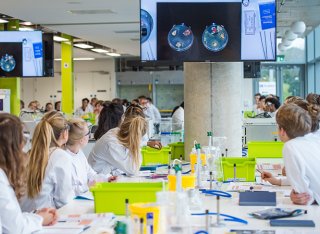 	Group of biosciences students wearing lab coats watching an experiment performed with petri dishes on screen