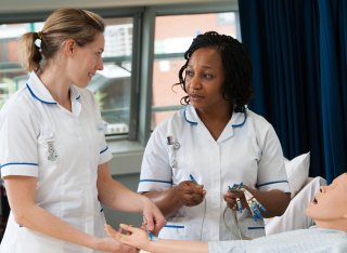 Two nursing students standing next to a mannequin laying on a bed