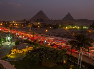 Time lapse photo showing the lights from moving traffic along a busy Cairo street with pyramids silhouetted against the skyline in the distance