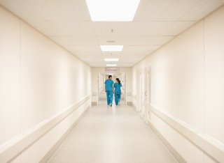 Two people wearing green scrubs can be seen a long way down a bright, white hospital corridor