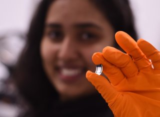 A blurred face smiles behind a hand, clad in an orange lab glove, holds a tiny rectangle of the new x-ray