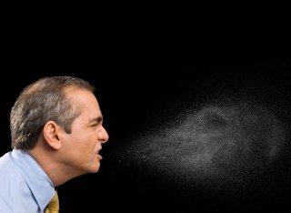 A middle aged white man wearing shirt and tie coughs. Against the plain black background, the spray from his mouth can be seen