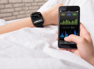 Two hands of a person lying in a bed with crisp white sheets. The left wrist wears a smart watch showing health data. The right hand holds a mobile phone