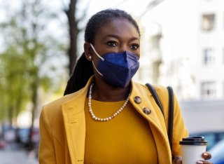 A commuting woman wearing a navy FFP2 face mask, yellow suit and pearl necklace carries a coffee along an urban street