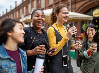 Four young women of different ethnic backgrounds enjoy a city break together. They are carrying phones and hot drinks and look as though they are having fun