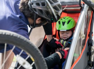 A dad wearing a silver bike helmet adjusts the straps of a toddler wearing a green bike helmet as he sits in a bike trailer