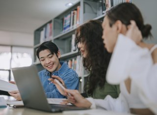 3 Asian LGBTQ+ adult students studying in college library writing notes and using laptop discussion on homework school project