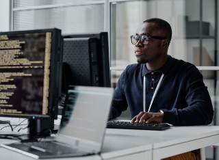 African American young developer in eyeglasses concentrating on his online work on computer sitting at workplace