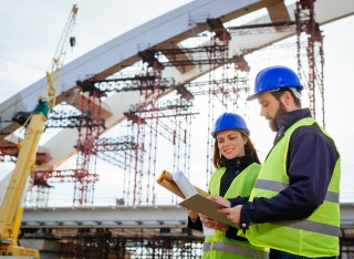 Two engineers, male and female, in protective helmets and reflective clothing, standing on construction site where large arch bridge is being built.