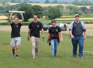 Four members of the Peryton Space team walk across a field, one holding their small rocket above his head