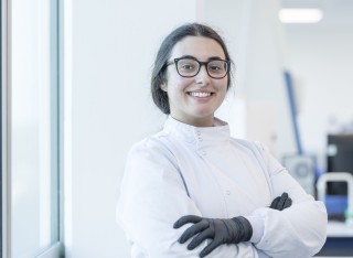 A biosciences student wearing glasses smiles confidently at the camera with her arms crossed.