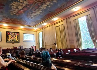 People sit in rows in a large, ornate room with projectors on the wall.