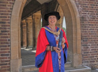 Dr Nicky Fox stands in the archways outside Guildford Cathedral holding her graduation scroll and wearing her graduation robes and cap. She is smiling at the camera