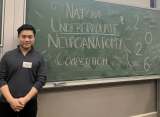 Michael stands in front of a blackboard that says 'National Undergraduate Neuroanatomy Competition'.