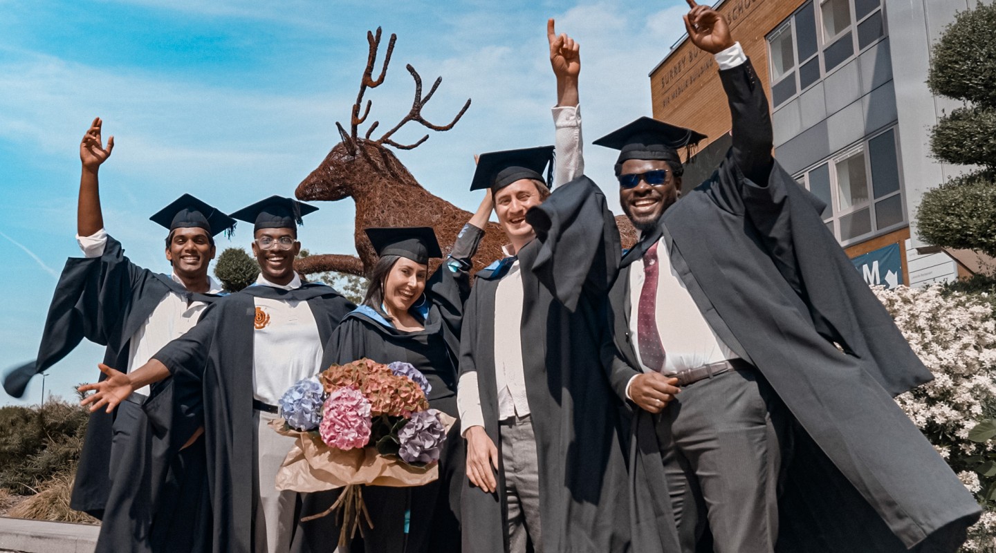 A group of postgraduate students at graduation cheering in front of a blue sky.