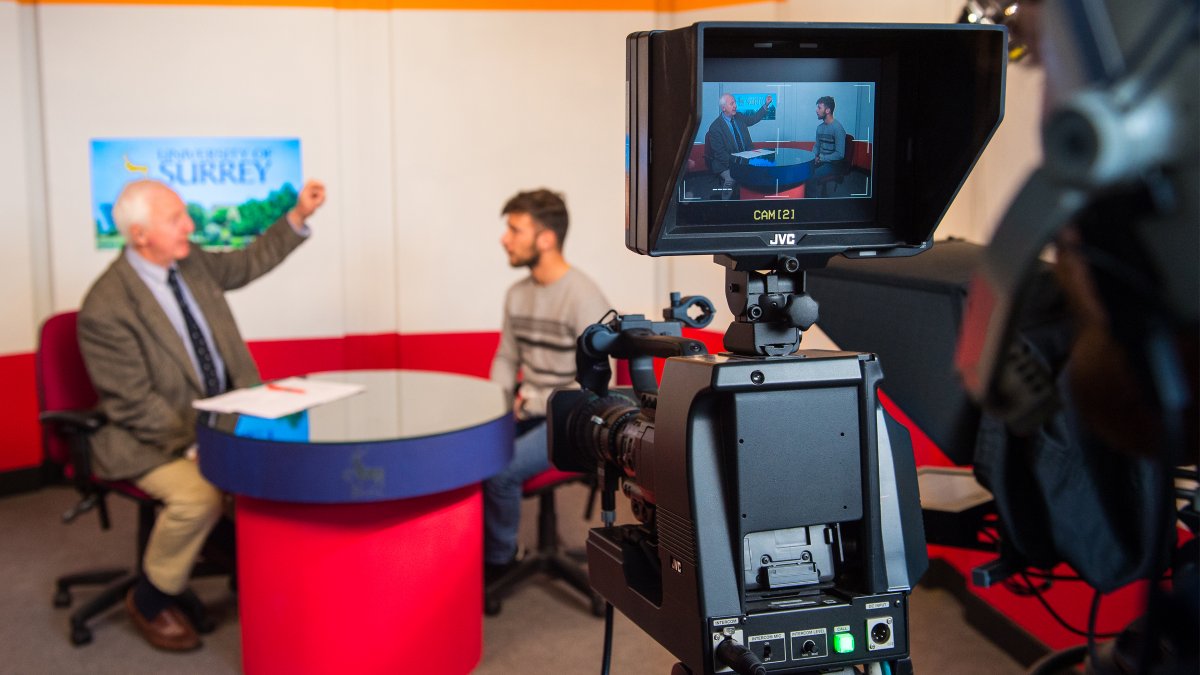 Camera set up for an interview with two males in background sitting on chairs