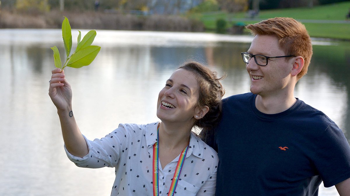 Two students are standing in front of the campus lake, one student is holding up a leaf which they are both looking at