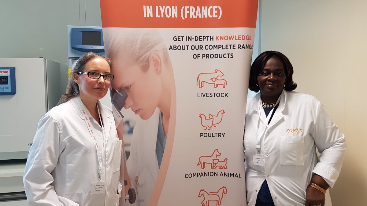 Georgina Cherry and Ruth Alafiatayo standing in front of a banner in Zoetis' diagnostic lab training centre in Lyon