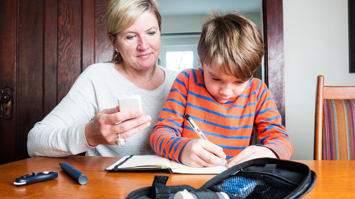 Mum watching her son write down his blood glucose levels in a notebook