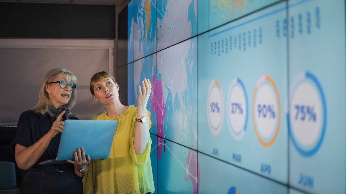 Two women discussing data on a giant screen to represent knowledge exchange