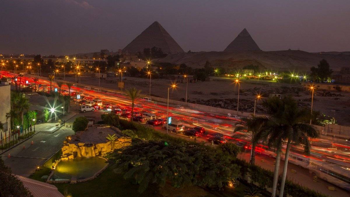 Time lapse photo showing the lights from moving traffic along a busy Cairo street with pyramids silhouetted against the skyline in the distance